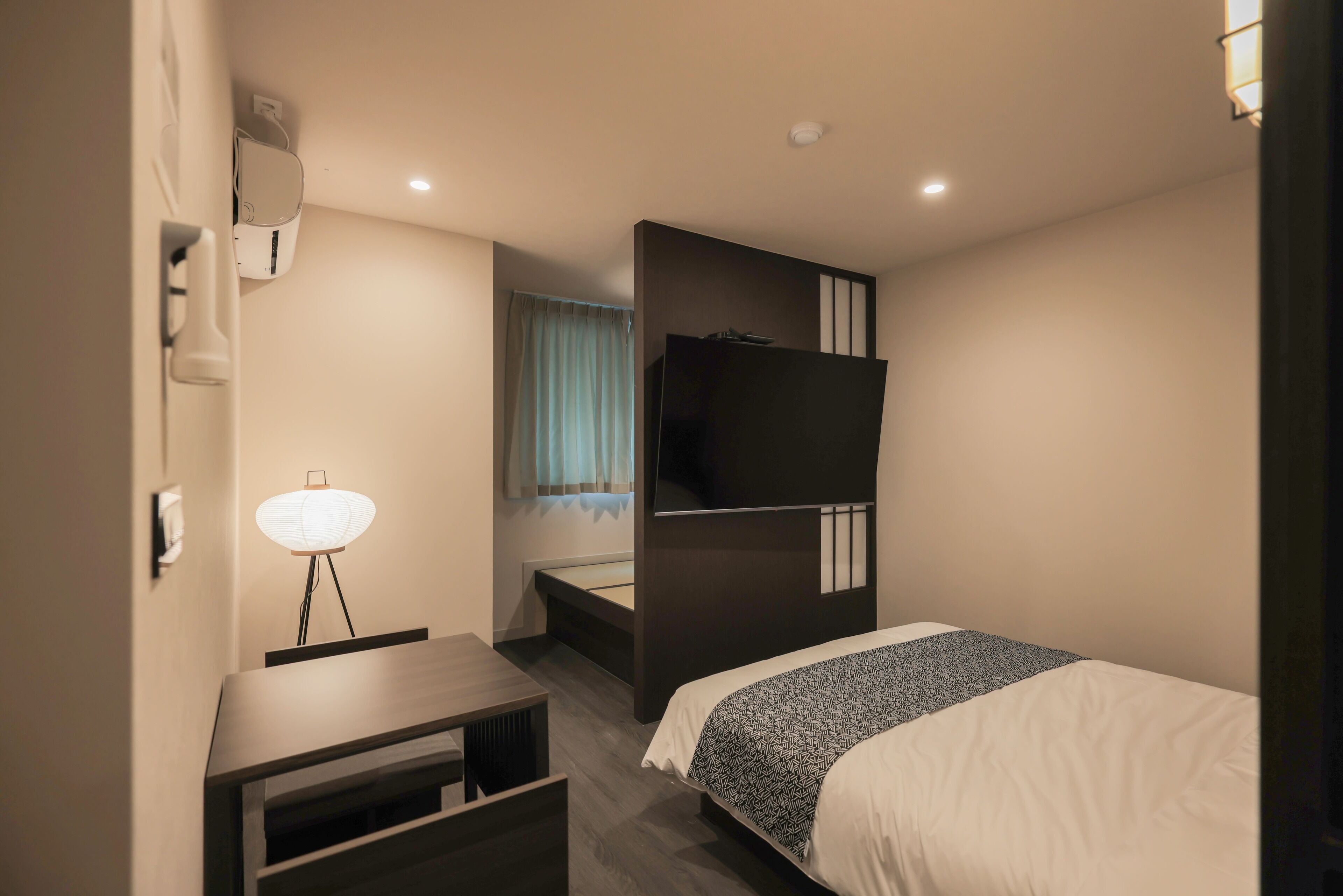 Moderni hotellihuone, jossa on sänky, työpöytä, tuoli, taulutelevisio ja ikkuna, jossa on verhot.
