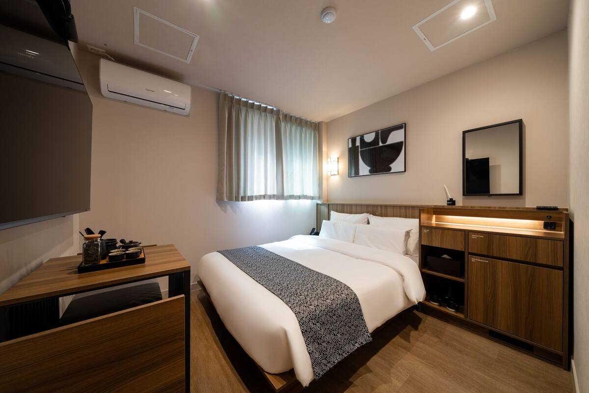 A modern hotel room with a large bed, bedside tables, a flat-screen TV, an air conditioning unit, a window with curtains, and a wooden cabinet with built-in lighting.