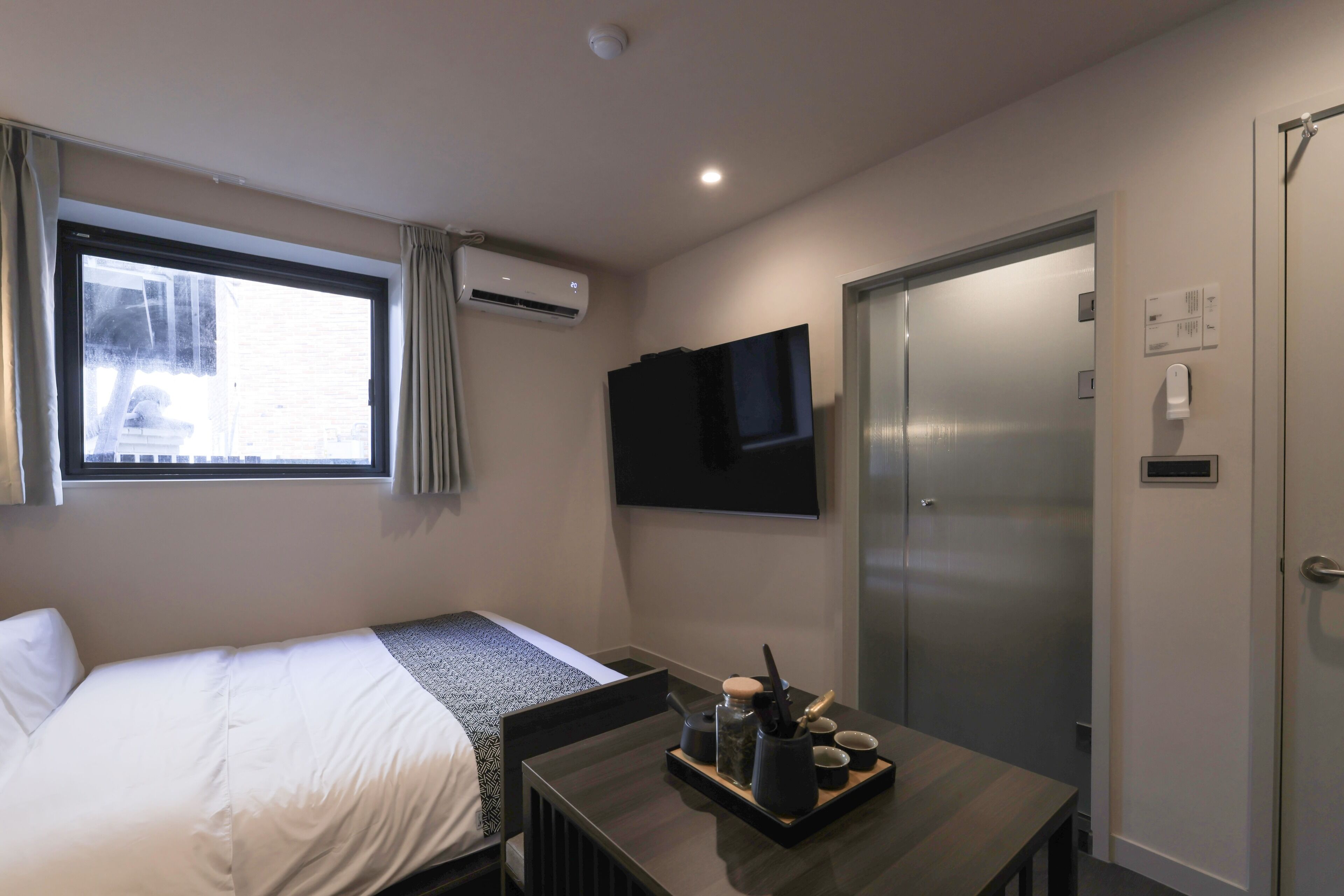 A hotel room with a bed, a desk with a tray of items, a TV mounted on the wall, and a door with a handle.