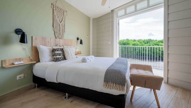 Ein Schlafzimmer mit Bett, Nachttisch, Lampe, Deckenventilator und Blick ins Grüne.