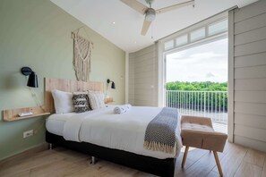 A bedroom with a bed, a bedside table, a lamp, a ceiling fan, and a view of greenery outside.