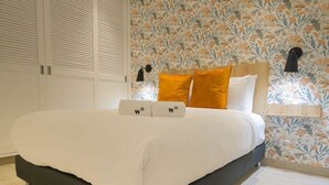 Un lit bien fait, avec une literie blanche et deux oreillers, une tête de lit en bois et un papier peint à motifs floraux.