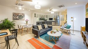 Un salon moderne avec un canapé, une table basse et un coin repas. On aperçoit une cuisine à l’arrière-plan, avec des appareils électroménagers et des placards.