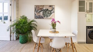Une cuisine moderne avec des meubles blancs, une table à manger entourée de chaises blanches, une plante en pot et un tableau accroché au mur.
