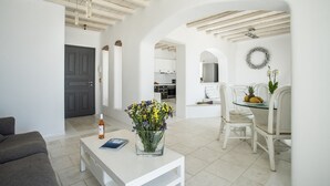 A spacious living area with a white sofa, a coffee table with a vase of flowers, and a dining table with chairs. There is a kitchen in the background.