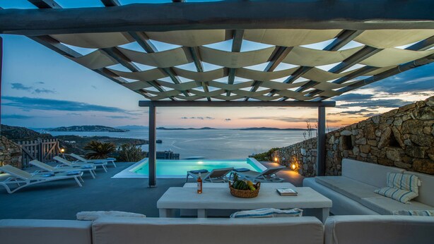 A poolside area with lounge chairs, a table with a basket of fruit, and a view of the sea at sunset.