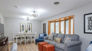 Ein modernes Wohnzimmer mit einem grauen Sofa, Poufs, einem Esstisch mit Stühlen und einem Fernseher.