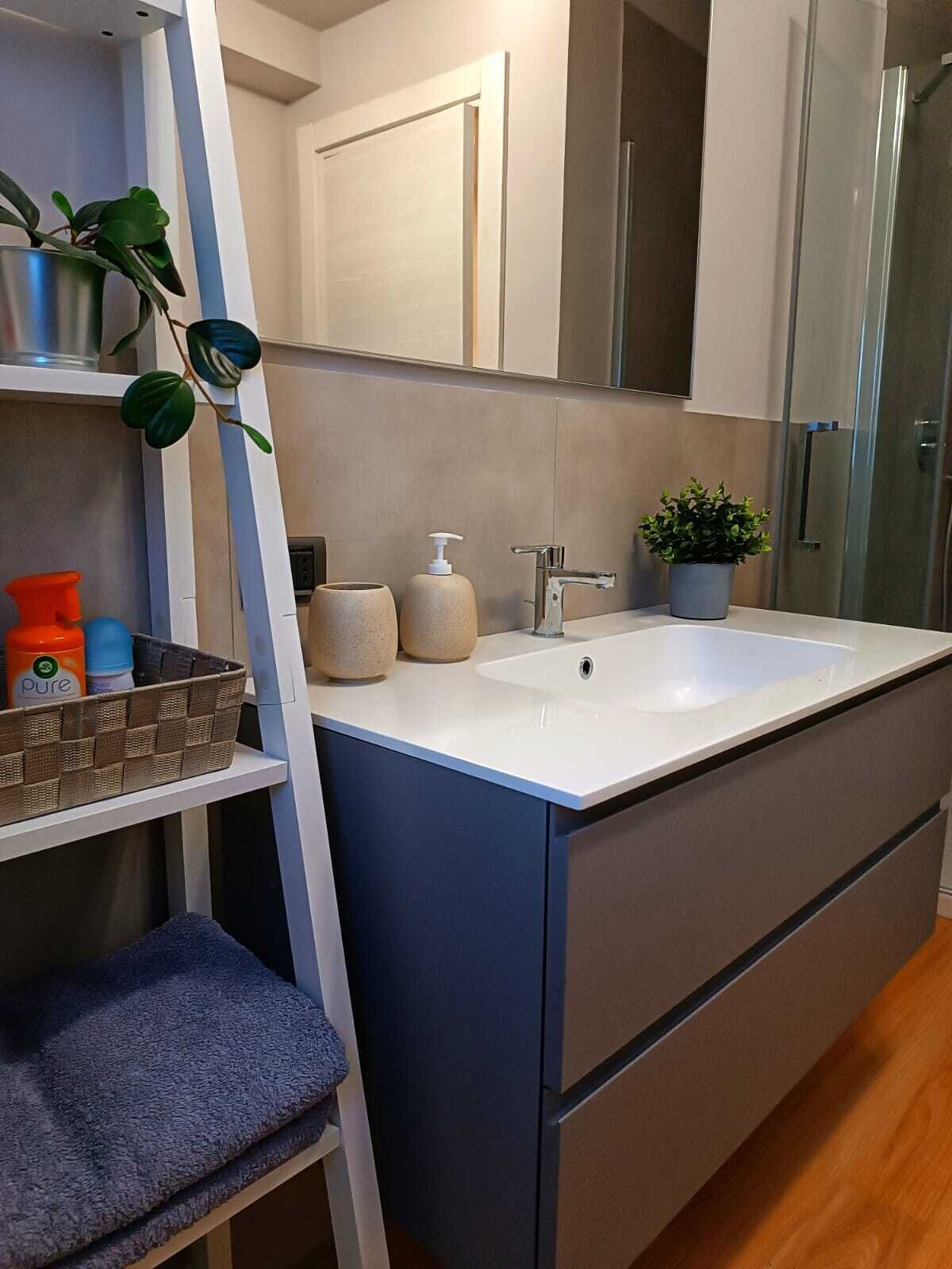 Et moderne badeværelse med en hvid vask, lysegrå skabe, en glasbrusekabine og en hylde med toiletartikler og en plante.