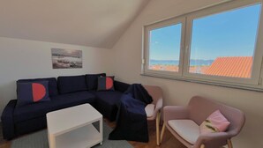 Een gezellige woonkamer met een donkerblauwe modulaire bank, een witte salontafel en een roze fauteuil. Een groot raam biedt uitzicht op een stadsbeeld.