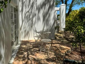 Une terrasse avec une table et des chaises en métal, un bâtiment blanc doté d’une fenêtre, et un arbre projetant son ombre sur le sol.