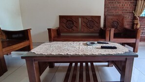 Eine hölzerne Bank mit aufwendigen Schnitzereien, ein Tisch mit Glasplatte und einem Tischtuch aus Spitze sowie zwei Fernbedienungen auf dem Tisch.