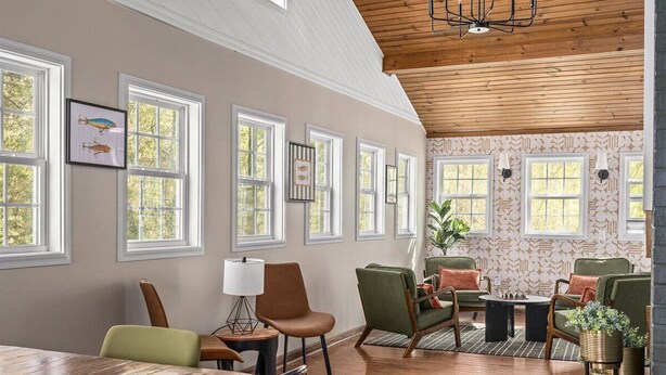 Una habitación amplia con techo alto, vigas de madera, ventanas grandes y una zona de estar acogedora con sillones tapizados en verde y una chimenea.
