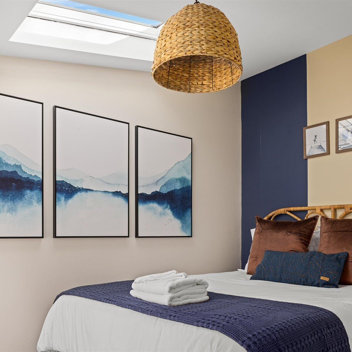Een slaapkamer met een bed, een geweven lamp en drie kunstwerken in lijsten aan de muur.