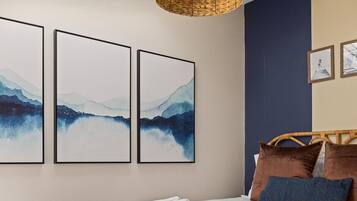 Een slaapkamer met een bed, een geweven lamp en drie kunstwerken in lijsten aan de muur.