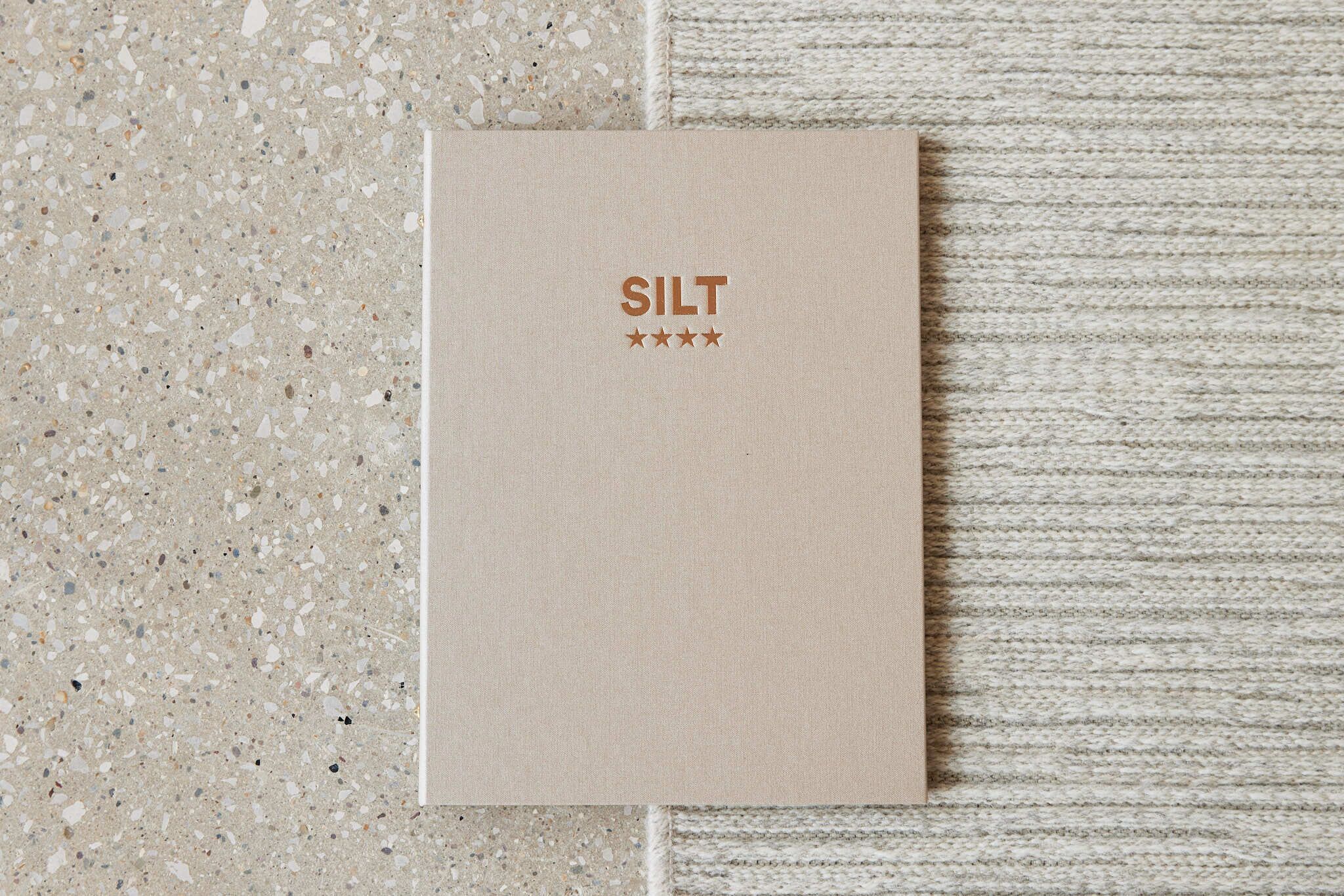 A booklet titled "SILT" with a textured cover, placed on a terrazzo floor next to a textured wall.