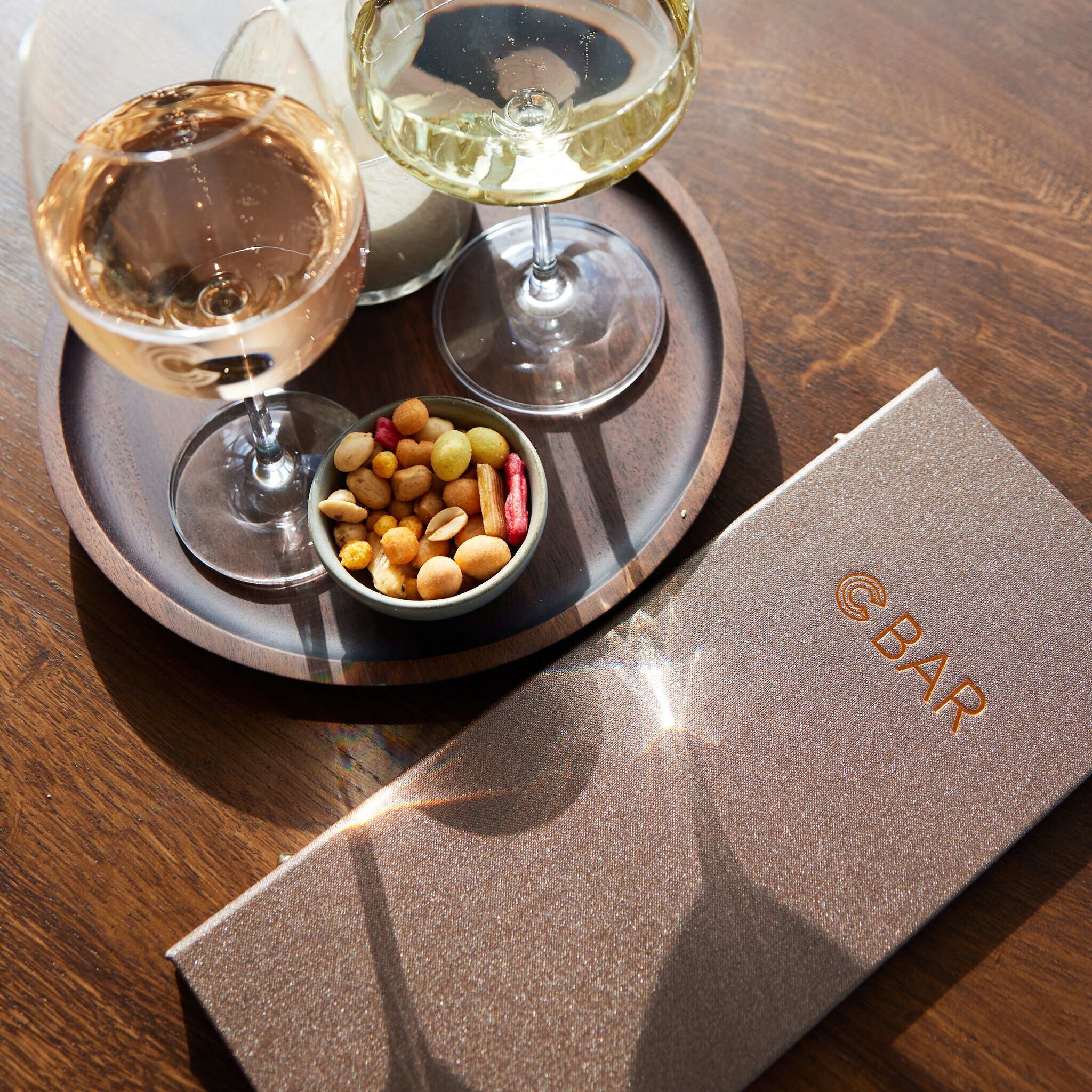 A tray with two glasses of white wine, a bowl of nuts, and a napkin with a logo.