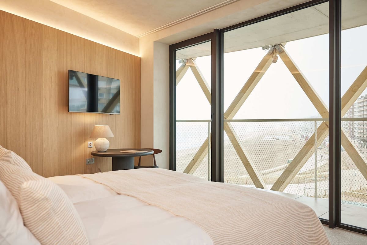 Ein Schlafzimmer mit einem großen Bett, einem Nachttisch inklusive Lampe, einem an der Wand montierten Flachbildfernseher und einem großen Fenster mit Blick auf ein Gebäude mit einer markanten, dreieckigen Dachkonstruktion.