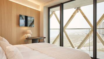 Een slaapkamer met een groot bed, een nachtkastje met een lamp, een flatscreen televisie aan de muur en een groot raam met uitzicht op een gebouw met een puntige dakconstructie.
