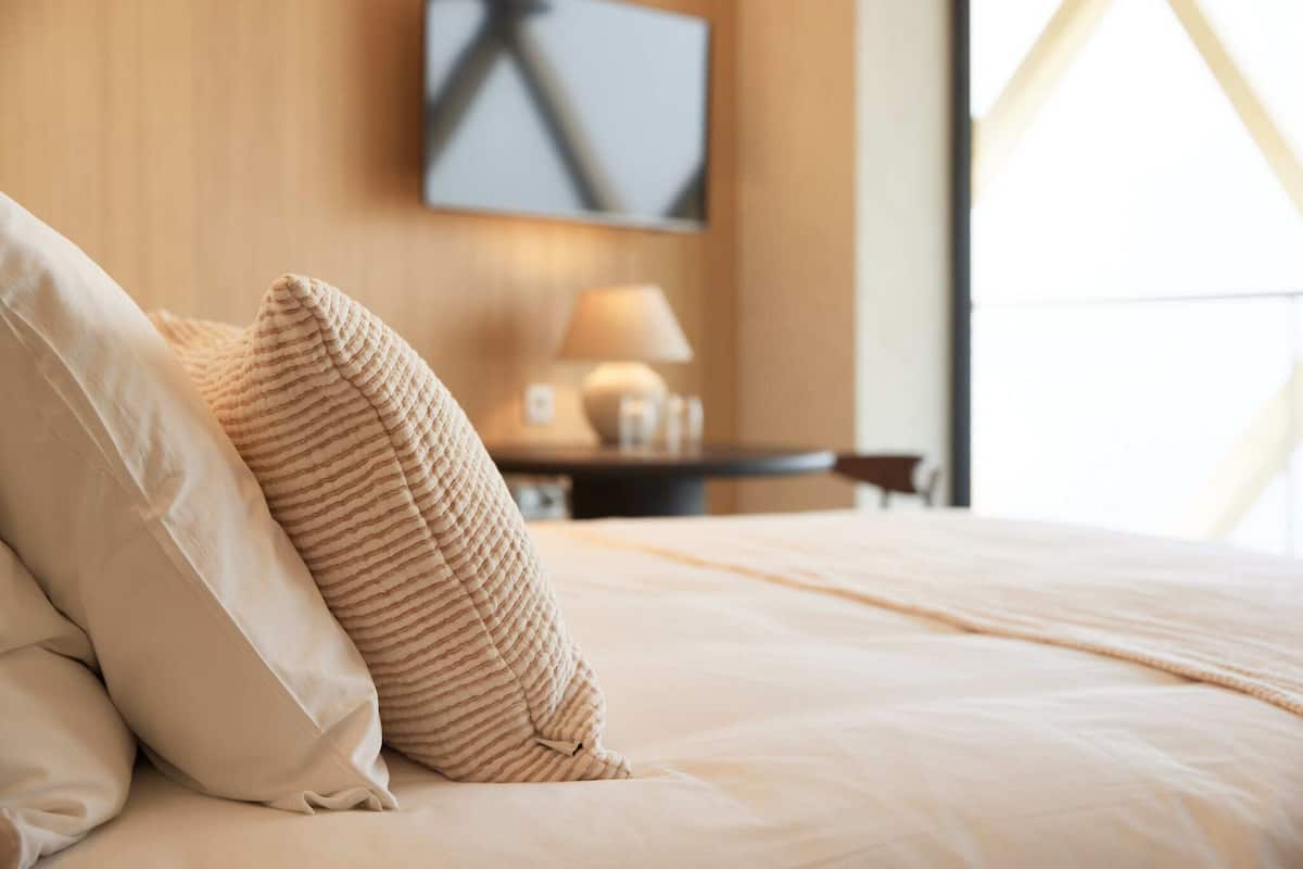 Ein ordentlich bezogenes Bett mit einem gestreiften Kopfkissen und einem schlichten Kopfkissen in einem Hotelzimmer mit einer Holzwand und einer Lampe auf einem Nachttisch.