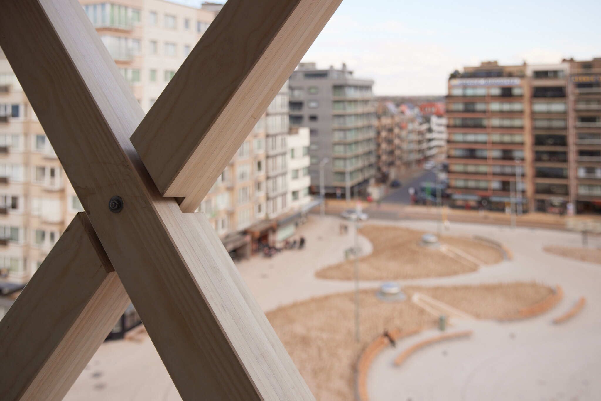 Uitzicht door een houten constructie, met een rond plein en meerhoogbouw op de achtergrond.