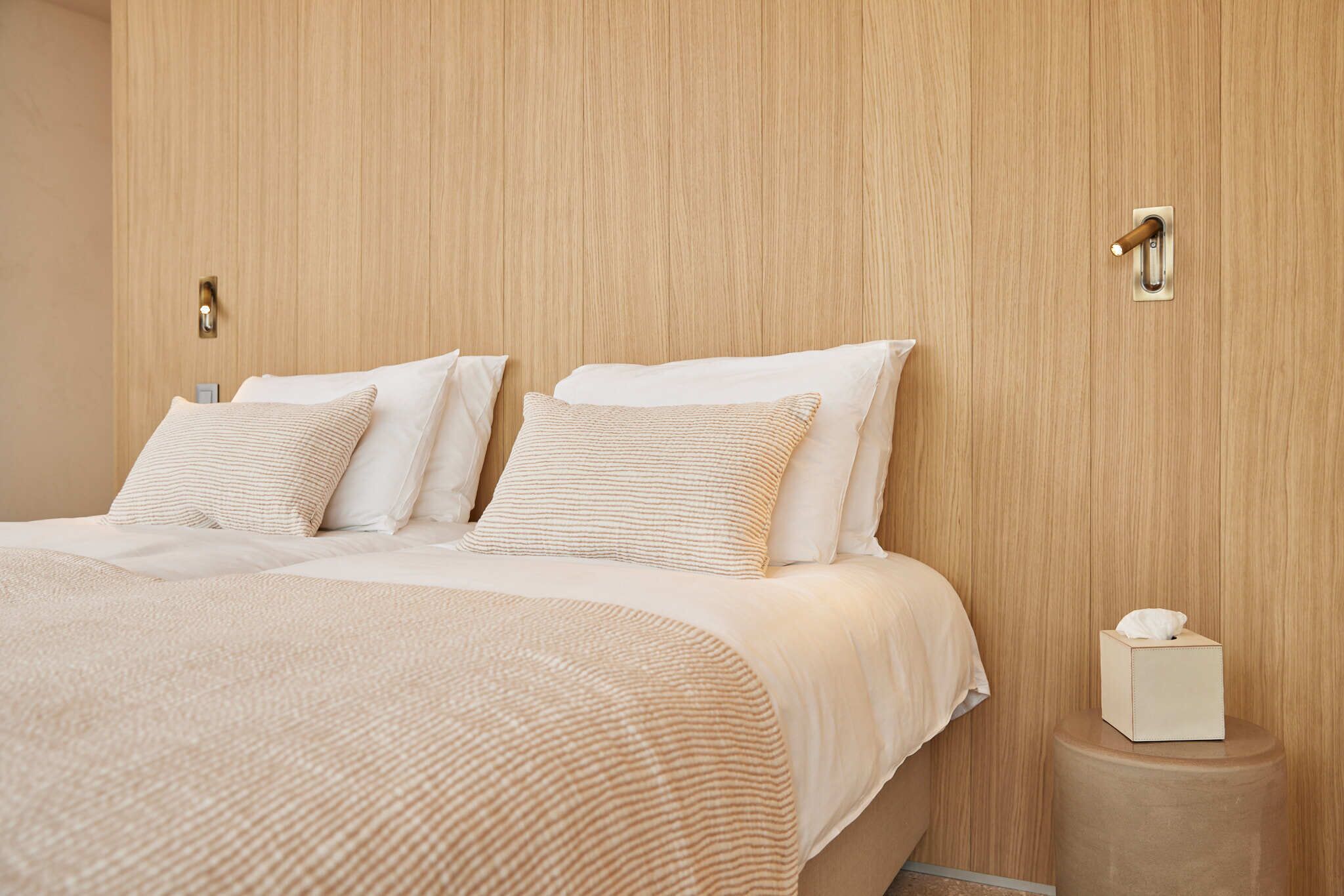 A neatly made bed with a beige comforter and pillows, a bedside table with a tissue box, and a wooden wall paneling.