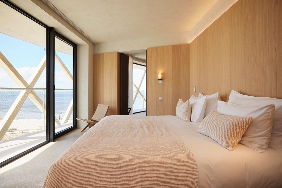 Ein modernes Hotelzimmer mit einem großen Bett, einem Sessel und Meerblick durch raumhohe Fenster.