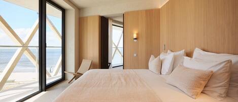 A modern hotel room with a large bed, a chair, and a view of the sea through floor-to-ceiling windows.