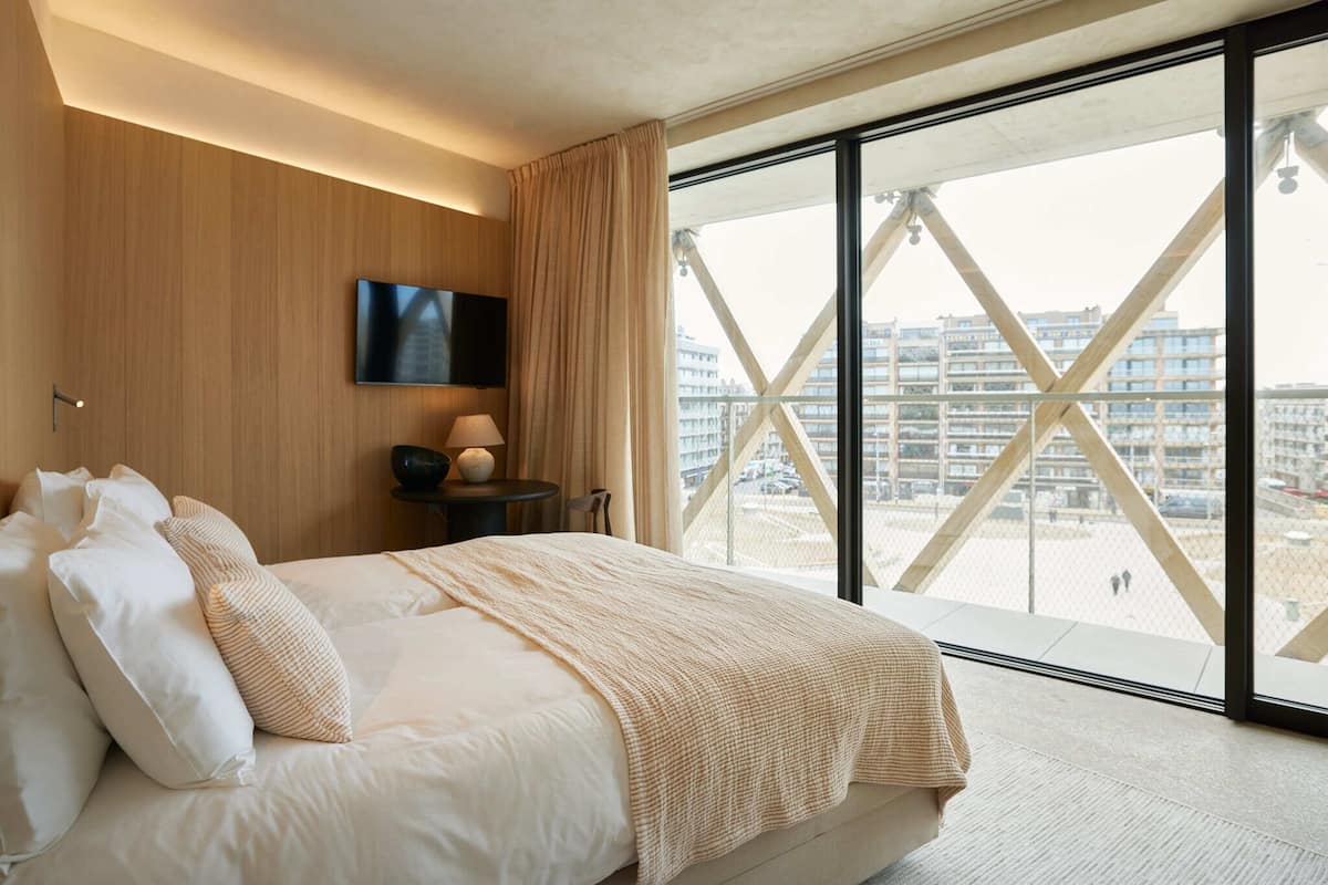 Ein modernes Hotelzimmer mit einem großen Bett, einem Flachbildfernseher und Blick auf eine Stadtkulisse durch raumhohe Fenster.