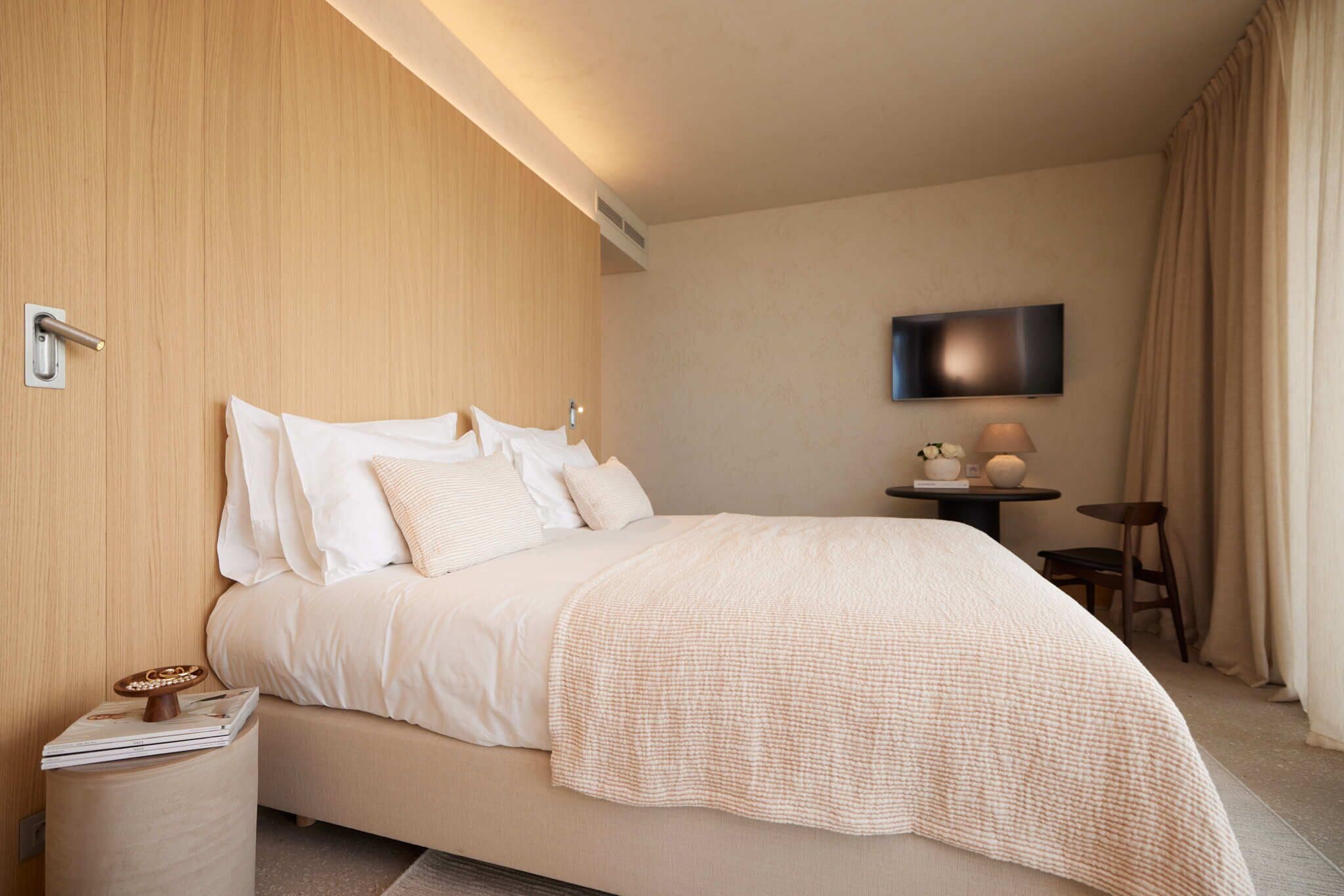 A hotel room with a bed, a bedside table, a lamp, a TV, and a small table with decorative items.