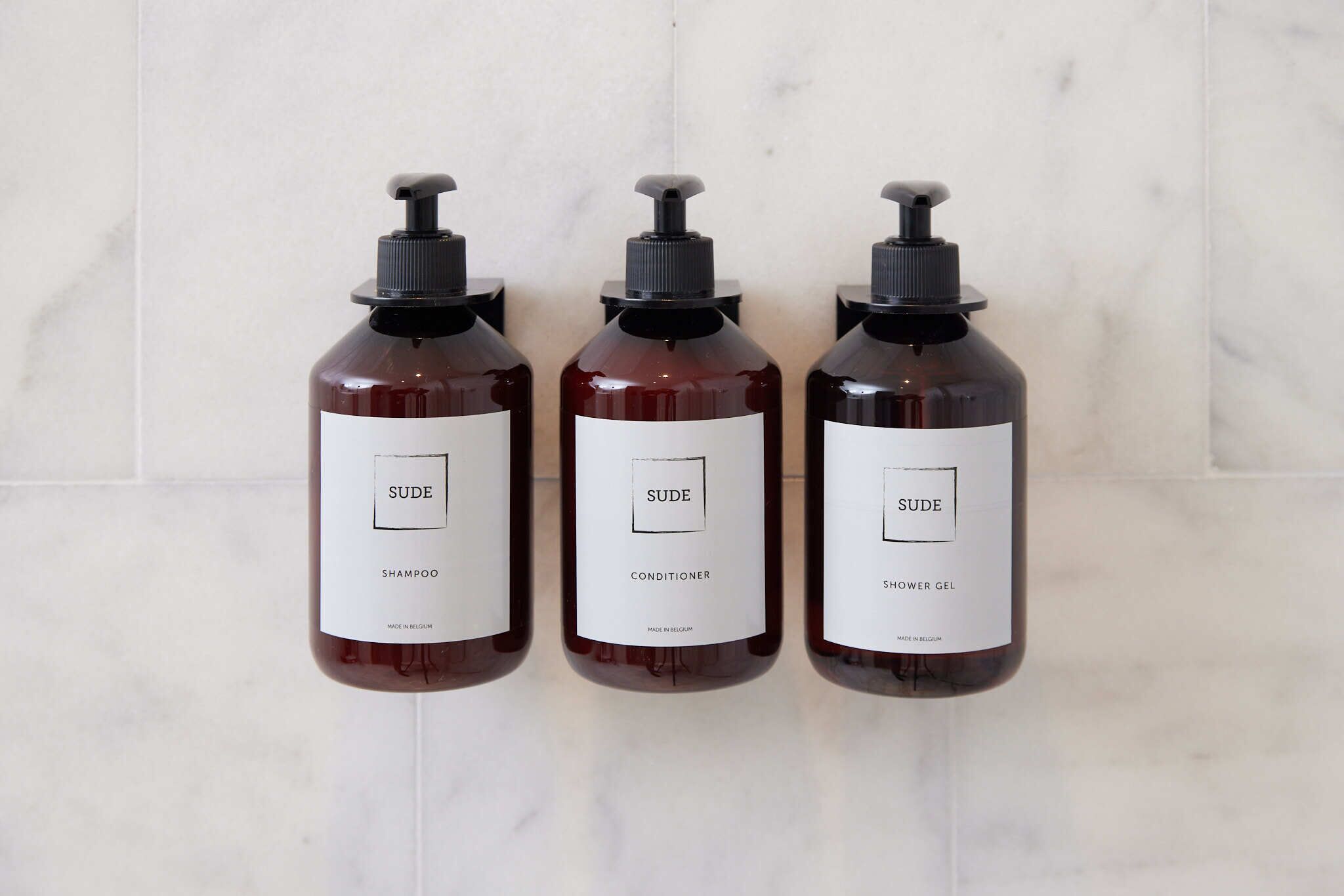 Three bottles of SUDE products: shampoo, conditioner, and shower gel, all with black pumps and white labels, against a marble background.