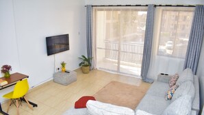 Un salón moderno con un sofá gris, un sillón amarillo, un televisor montado en la pared y un balcón con barandilla.