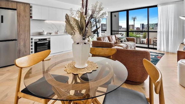 A modern living room with a glass dining table, brown leather sofa, and a view of the city through large windows. - Cityscape at The Elliott in the Heart of Hobart (Hobart)