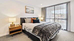 Una habitación de hotel moderna con una cama grande, una mesita de noche con lámpara, vistas de la ciudad a través de amplios ventanales y un cuadro en la pared.