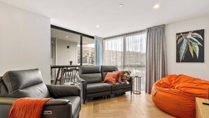 Una sala moderna con un sofá de cuero negro, un sillón gris y un asiento tipo puff naranja. Hay una zona de comedor con una mesita y sillas, y un ventanal con cortinas.