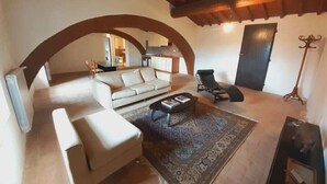 Un'ampia zona giorno con divano, tavolino e poltrona. Si nota un soffitto in legno e una porta sullo sfondo.