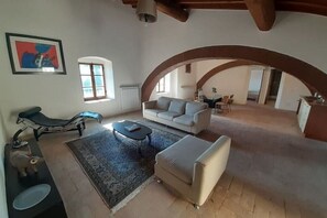 Un grand espace de vie comprenant un canapé, une table basse et une moto. Une fenêtre et une porte en arc donnent accès à une autre pièce.