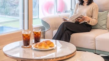 Una persona sentada en un sofá leyendo una revista, con una mesa frente a ella que tiene un plato de comida y dos vasos de té helado.