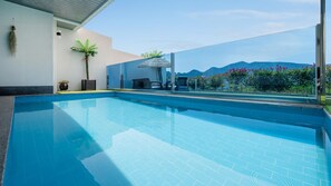 An indoor swimming pool with clear blue water, surrounded by a glass wall offering a view of mountains and flowers. - Namhae Eagless Poolvilla Pension (Namhae)