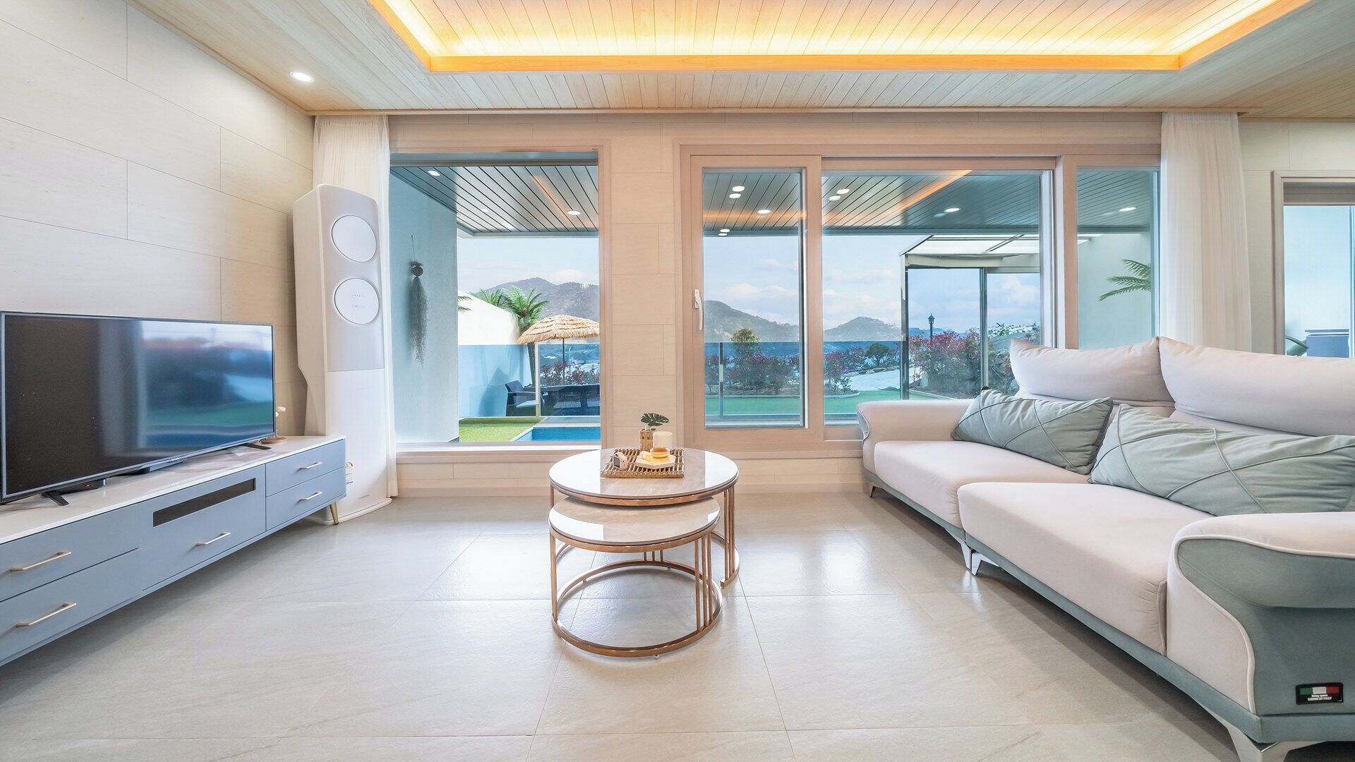 A modern living room with a flat-screen TV, a sofa, a coffee table, and large windows offering a view of a pool and mountains.