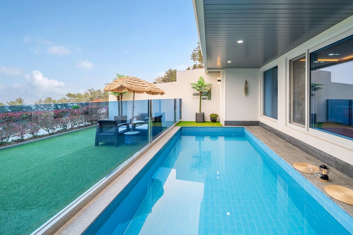 Una piscina con cerramiento de vidrio, una sombrilla de paja y mobiliario exterior.