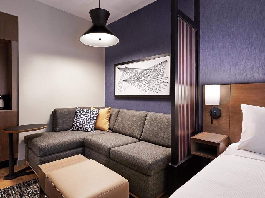 A modern hotel room with a sectional sofa, a bed, a bedside table, a coffee machine, and a wall-mounted lamp.