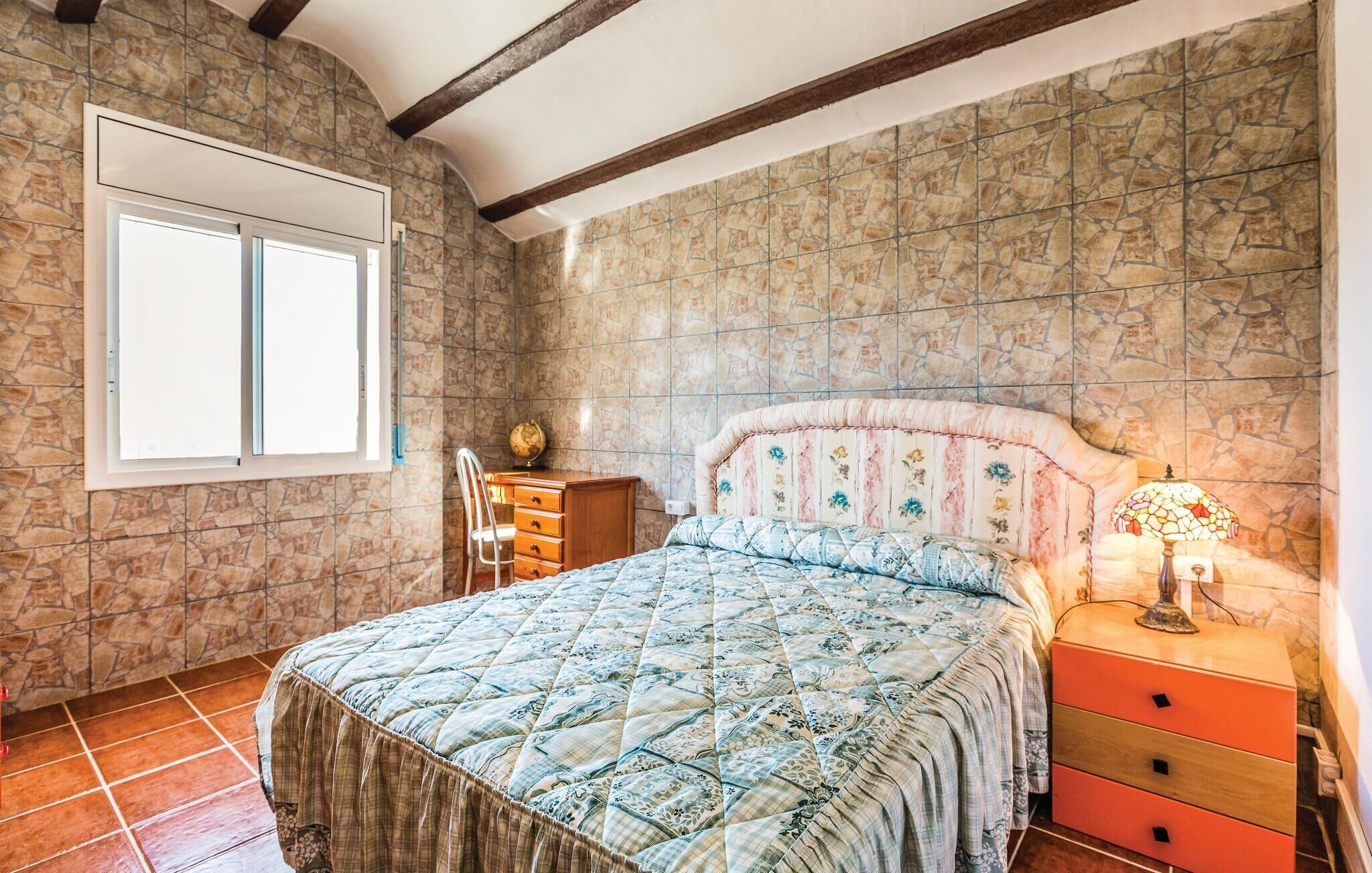 A bedroom with a stone wall, a bed with a floral headboard, a dresser, a chair, and a nightstand with a lamp.