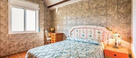 A bedroom with a stone wall, a bed with a floral headboard, a dresser, a chair, and a nightstand with a lamp.