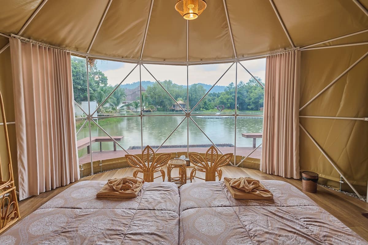 A tented room with a bed, two chairs, a table, and a view of a lake.