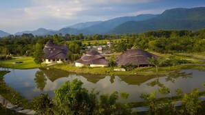 A traditional thatched-roof building with a pond in the foreground, surrounded by lush greenery and hills in the background. - Eco Eyes Village (Nakhon Nayok)