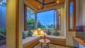 Una habitación acogedora con un ventilador de techo de madera, ventanas grandes con marcos de madera, un banco acolchado con almohadas, una mesita redonda con una lámpara y flores, y un televisor de pantalla plana sobre un soporte de madera.