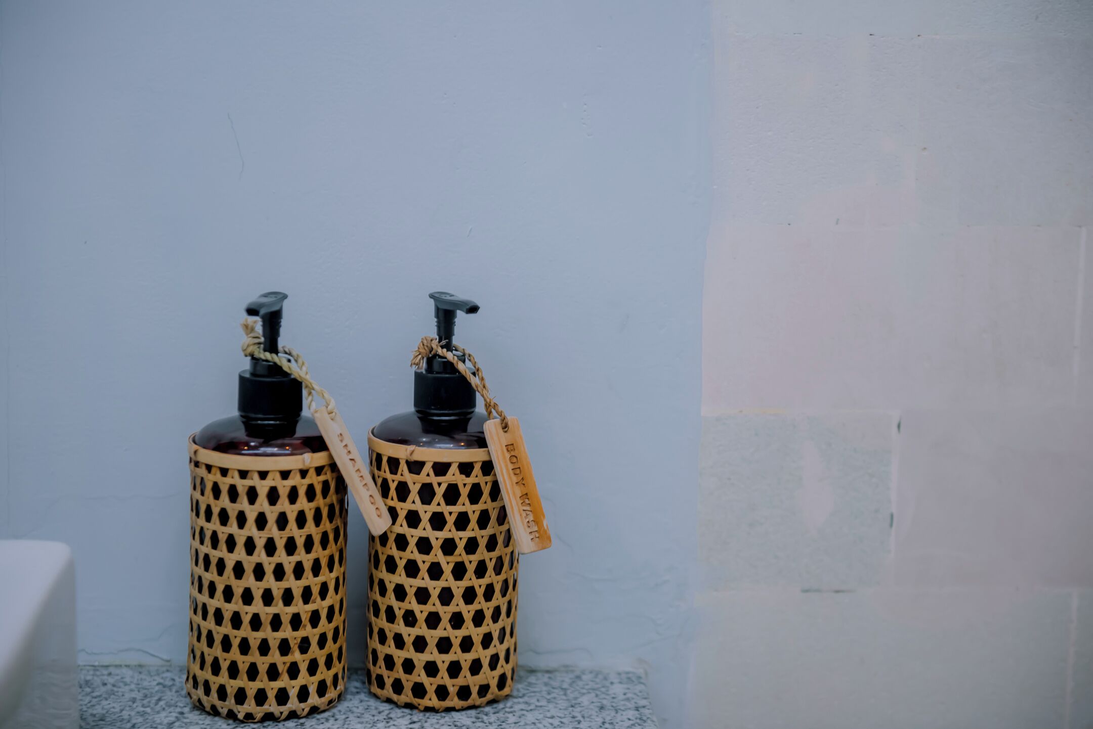 Two wicker dispensers with black pumps and tags, placed against a plain wall.