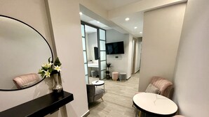 A modern hotel room with a large mirror, a small round table, two pink cushioned chairs, a TV mounted on the wall, and a visible bathroom area with a sink and mirror.