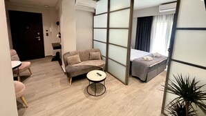 A modern hotel room with a sofa, a small round table, a bed with a grey duvet, and an air conditioning unit.