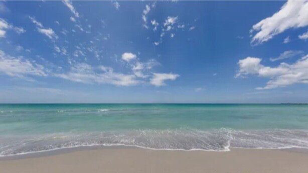 A beach with clear turquoise water and a sandy shore under a blue sky with scattered clouds.